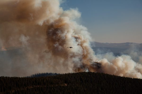 A helicopter flying against a backdrop of smoke with trees below.